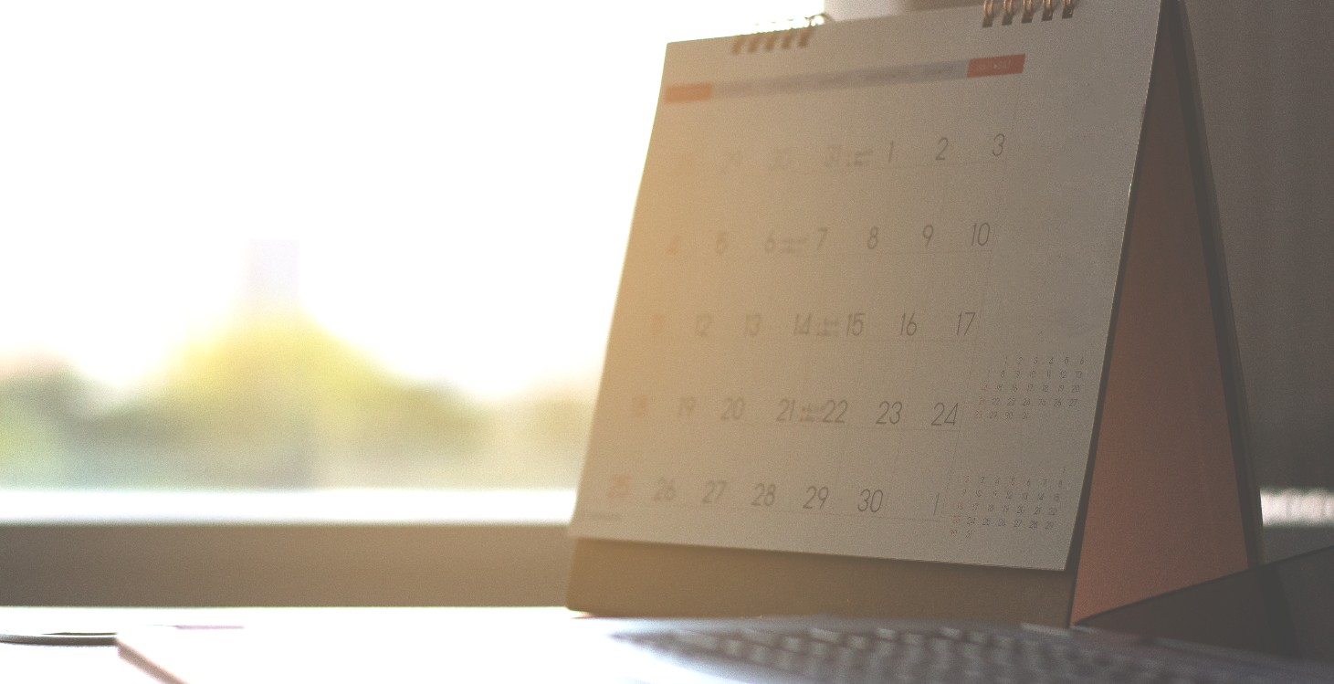 Folded monthly calendar on a desk with an open laptop next to it, in the background there is evening sunlight coming through the window. 