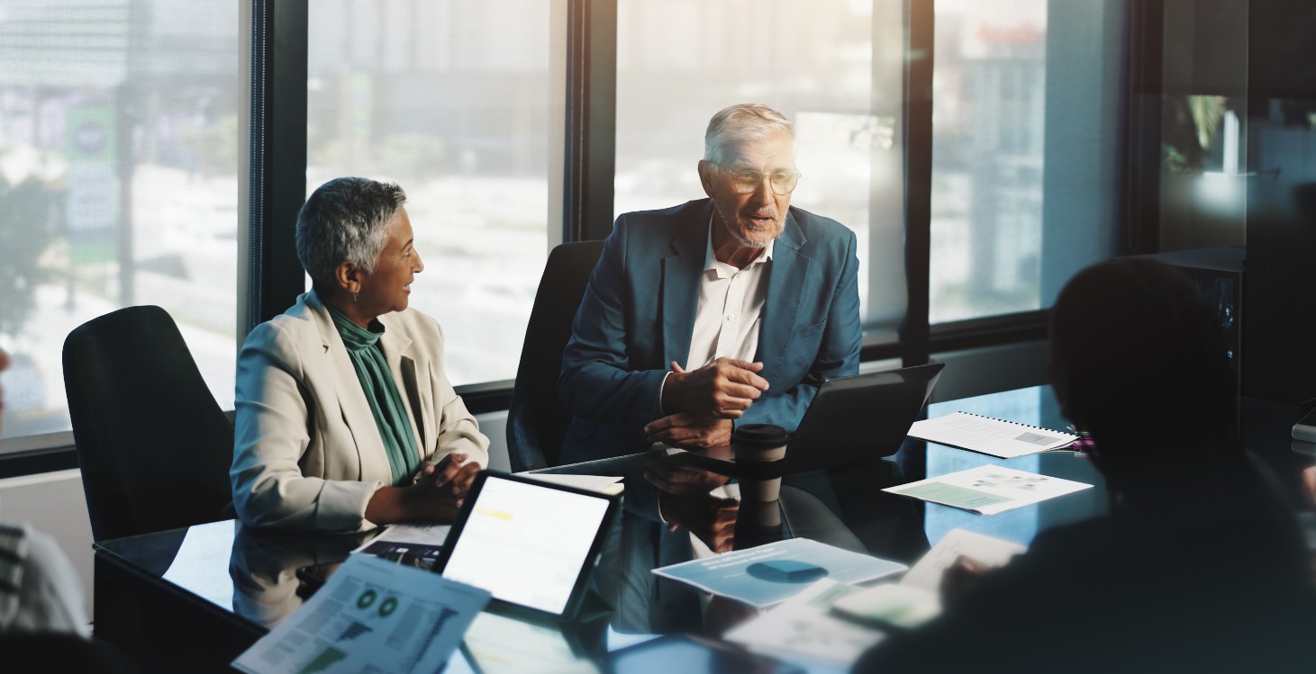 Two older business professionals in a meeting with other professionals sitting around a meeting table