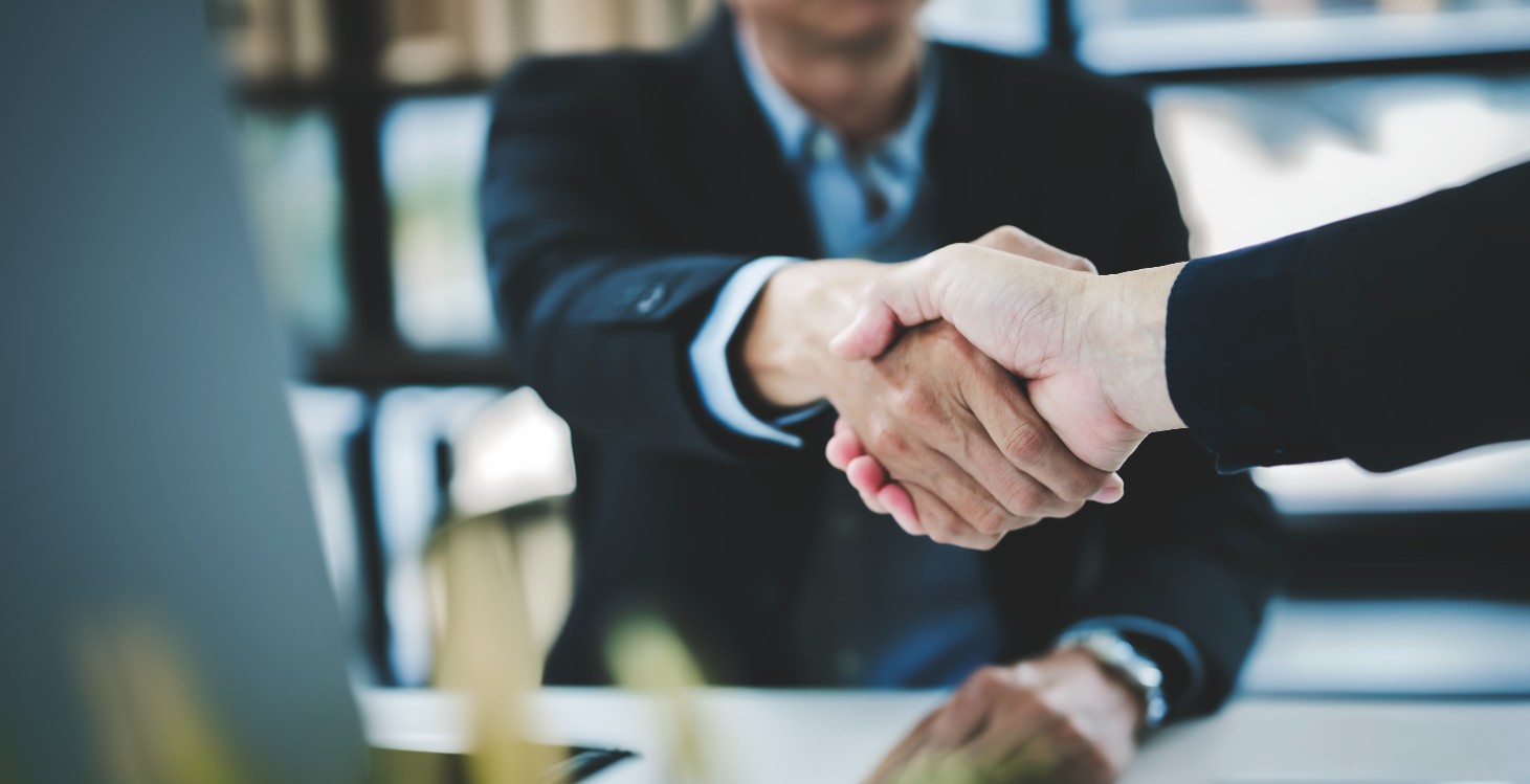 Two people in business attire shaking hands across a desk, suggesting a professional agreement or partnership.