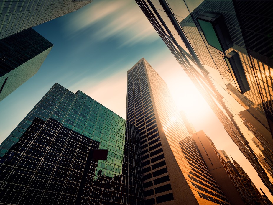 A wide, low-angle panoramic view of tall glass skyscrapers in a city during sunset, featuring warm golden reflections and cool teal shadows.